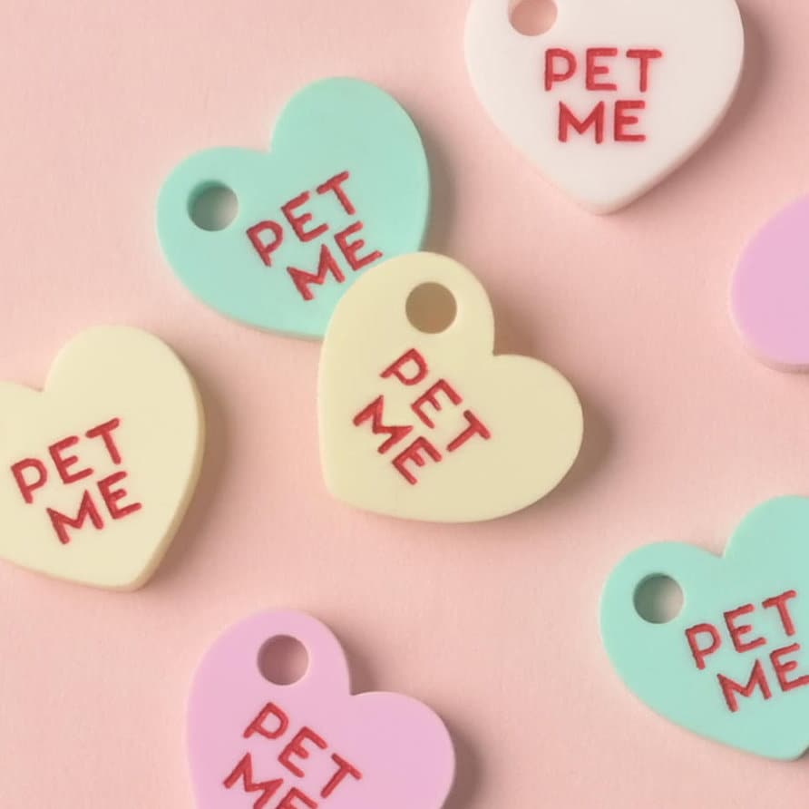 A close-up cluster of six small, heart-shaped acrylic mini-charms, each engraved with the red slogan 'PET ME.' The charms are in assorted pastel colors: white, mint green, light yellow, and lilac purple, displayed on a solid pink background. Small Pet Me Heart, Candy Inspired, Valentine's Day, Seasonal Mini, Tiny Accessory for Pet ID Tag, Cats and Dogs Add-on Charm