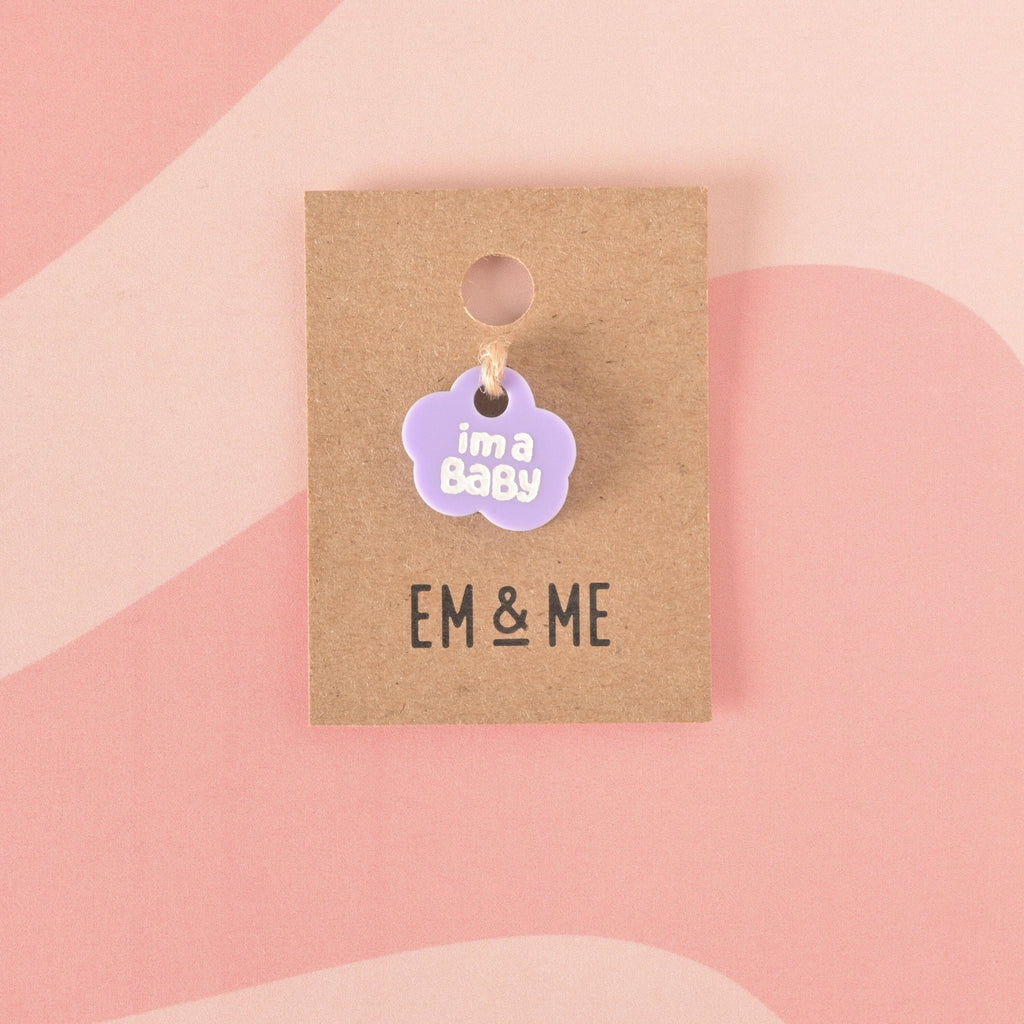 A close-up of a small, light purple cloud-shaped charm that says "i'm a Baby" in white letters on a brown kraft paper card on wavy pink background