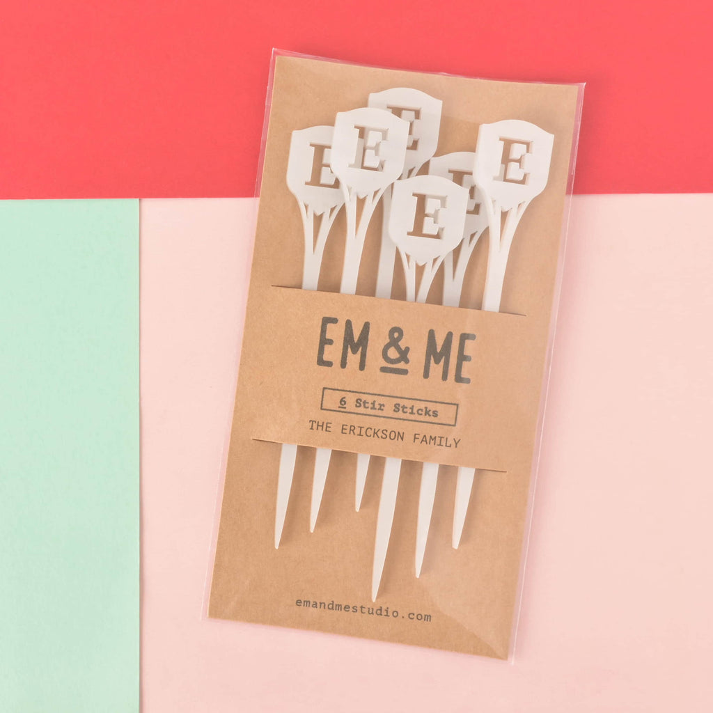 A packaged set of six white monogram cocktail stirrers, featuring the letter 'E' in a shield shape at the top. The packaging is a brown card that reads "EM & ME," "6 Stir Sticks," and "THE ERICKSON FAMILY." The item is displayed on a background of solid pink, mint green, and red.
