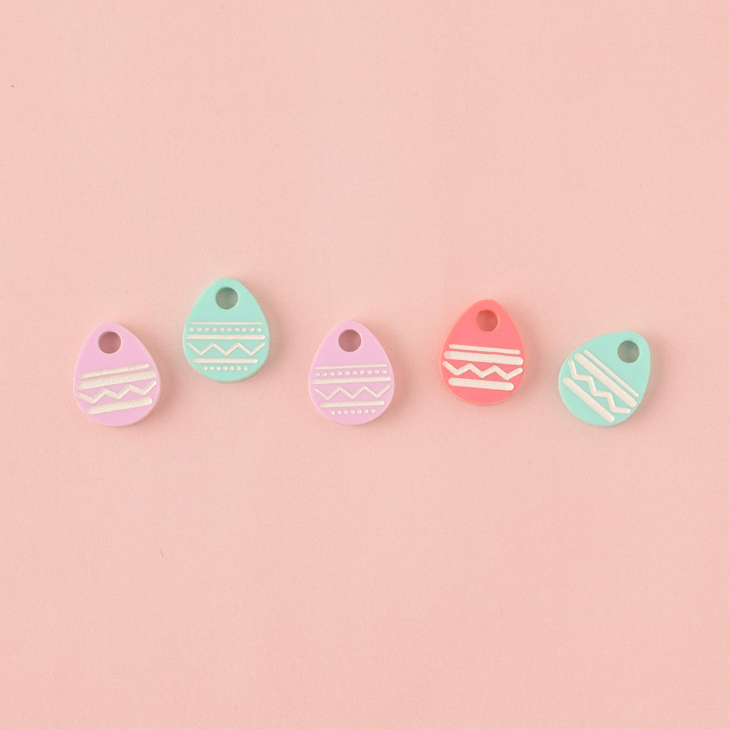Five small acrylic charms shaped like Easter eggs, engraved with white zigzag and stripe patterns. The charms are in pastel colors: pink, light purple, and mint green, and are arranged horizontally on a solid light pink background.