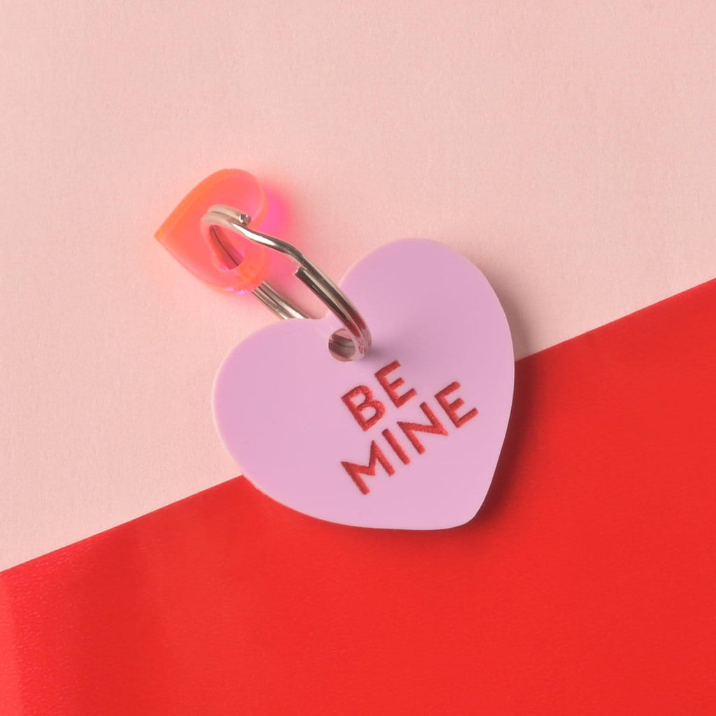 Close-up of a single light purple (lilac) heart-shaped acrylic pet ID tag, engraved with the darker red slogan 'BE MINE.' A translucent orange/pink heart charm is attached to the silver split ring. The tag is displayed on a color-blocked background of light pink and bright red. Conversation Hearts, Valentines Heart Dog tag, Customize front and back, ID tag for cats and dogs, Personalized Pet Tag