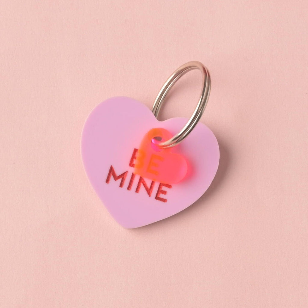 Close-up of a single light lavender pink heart-shaped acrylic pet ID tag, engraved with the darker pink slogan 'BE MINE.' A small, layered pink and orange heart charm is attached to the silver split ring, displayed on a solid pink background.Conversation Hearts, Valentines Heart Dog tag, Customize front and back, ID tag for cats and dogs, Personalized Pet Tag