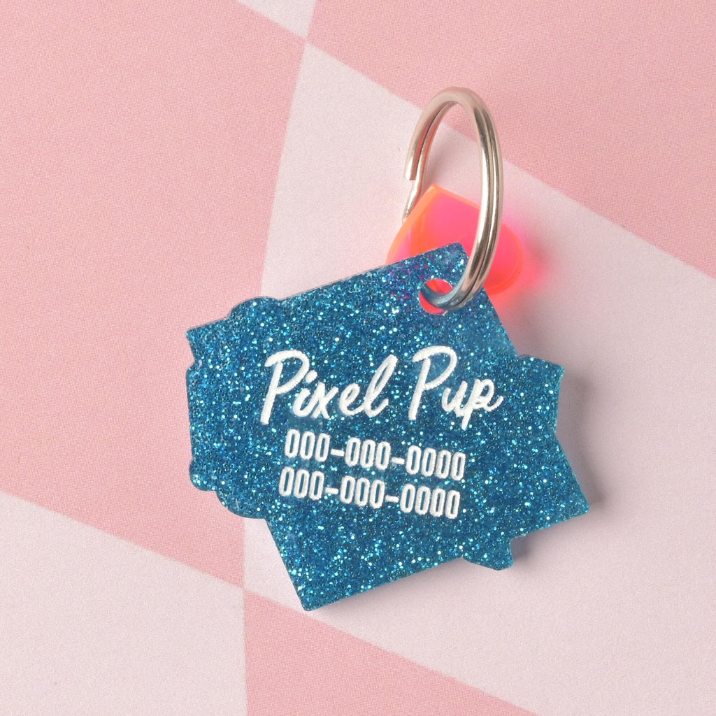 A custom, shield-shaped pet ID tag made from vibrant blue glitter acrylic. The tag is engraved with the name 'Pixel Pup' and three lines of phone numbers in white, and accented with a small iridescent pink/orange charm.