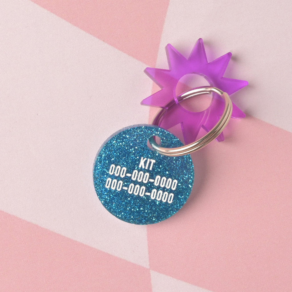 A custom, circular pet ID tag made from vibrant blue glitter acrylic. The tag is engraved with the name 'KIT' and phone numbers, and accented with a translucent purple sunburst charm on a split ring. Personalized Barbie-Inspired Cat Tag, Custom Engraved with Contact Info, Limited Edition "Actually Purrfect" Design, Dollhouse for kittens