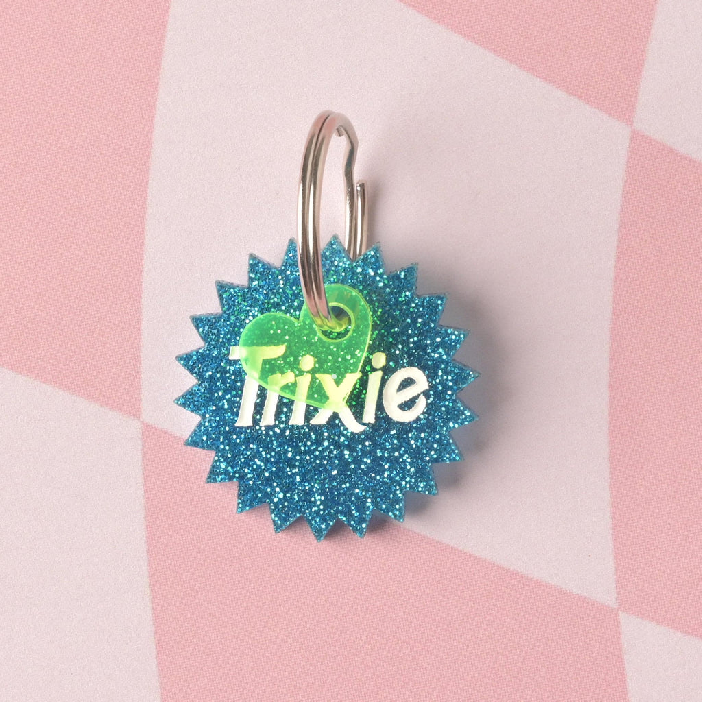  A custom pet ID tag shaped like a starburst in vibrant blue glitter acrylic. The tag is engraved with the name 'Trixie' and accented with a small neon green heart charm on a split ring, set against a pink checkered background. Barbie Inspired Trixie Personalized Pet Tag, Barbie Sunburst Pet Tag for Cats and Dogs, Limited Edition, Custom Name and Info Engraved