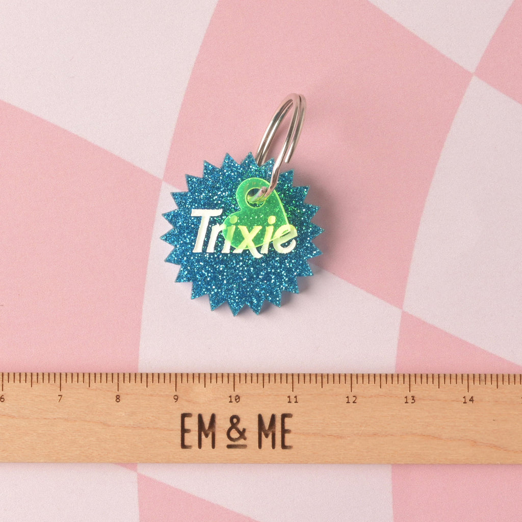  A custom pet ID tag shaped like a starburst in vibrant blue glitter acrylic. The tag is engraved with the name 'Trixie' and accented with a small neon green heart charm, shown above a wooden ruler for size reference. Barbie Inspired Trixie Personalized Pet Tag, Barbie Sunburst Pet Tag for Cats and Dogs, Limited Edition, Custom Name and Info Engraved