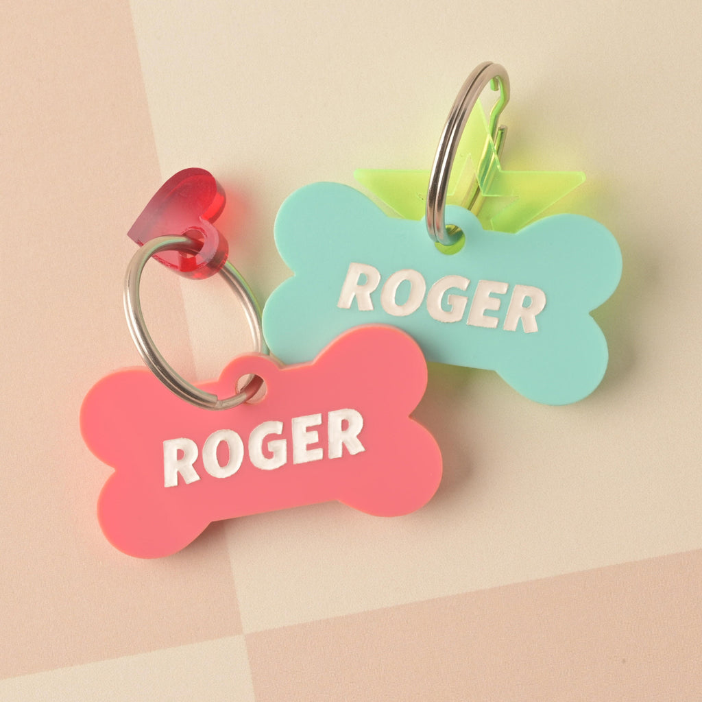 Two bone-shaped acrylic pet ID tags placed closely together. The front tag is pink, personalized with the name 'ROGER,' and has a small red heart charm. The back tag is mint green, also personalized with 'ROGER,' and has a neon star charm. Bones Day, Bone shaped Dog Tag, Personalized Pet Tag, Cat and Dog ID Tag