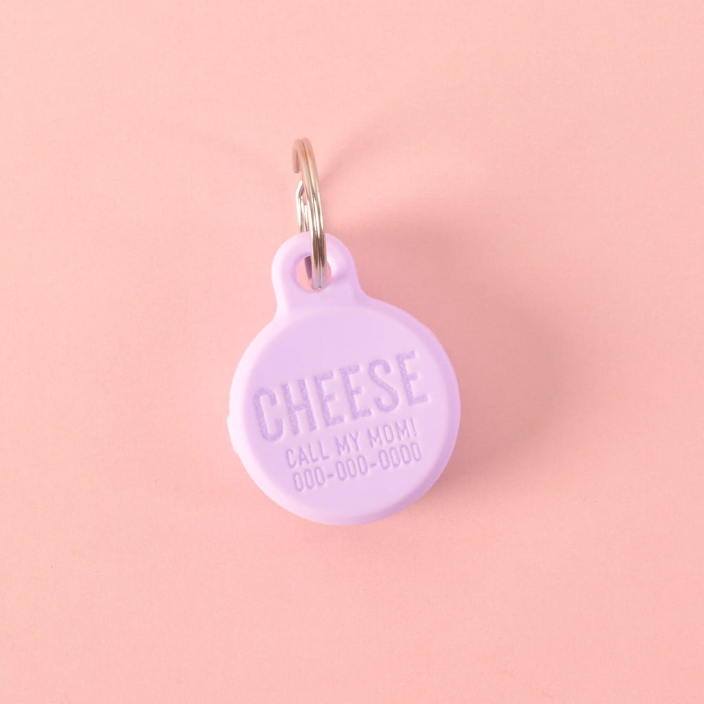 A personalized lavender round silicone pet tag that says "CHEESE and "CALL MY MOM!" with a placeholder phone number on it. The tag is attached to a silver ring, perfect for a collar, and is displayed on a solid pink background.