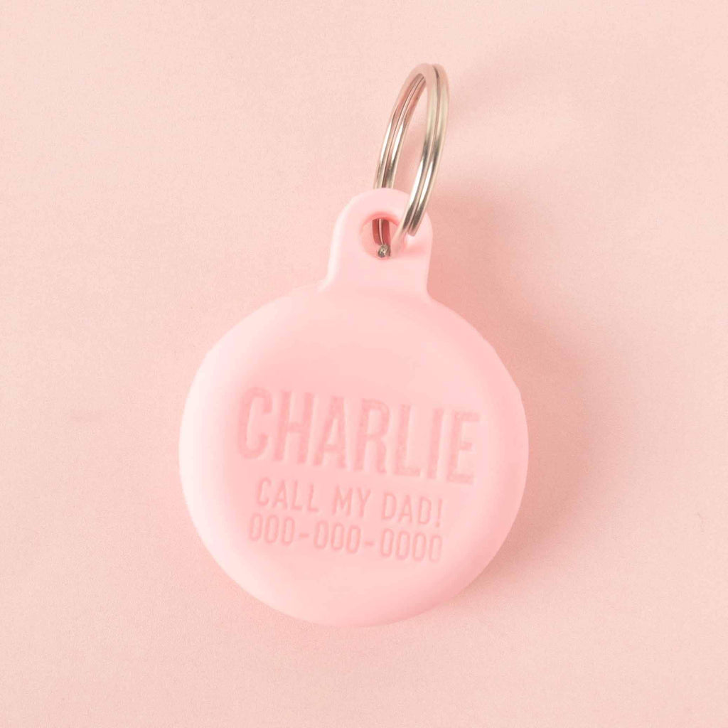 A personalized pink round pet tag that says "CHARLIE" and "CALL MY DAD!" with a placeholder phone number on it. The tag is attached to a silver ring, perfect for a collar, and is displayed on a solid pink background.