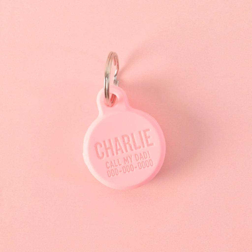 Personalized Engraved GPS Pet Tag Holder, SA personalized pink round pet tag that says "CHARLIE" and "CALL MY DAD!" with a placeholder phone number on it. The tag is attached to a silver ring, perfect for a collar, and is displayed on a solid pink background.