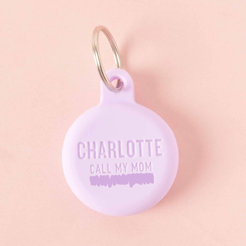 A personalized light purple round pet tag that says "CHARLOTTE" and "CALL MY MOM" with a placeholder phone number on it. The tag is attached to a silver ring, perfect for a collar, and is displayed on a solid pink background.