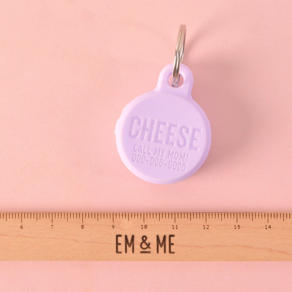 A personalized purple round silicone tag designed to hold a Tile tracker. The tag says "CHEESE" and has a placeholder phone number on it. A wooden ruler with the "EM & ME" logo is placed below the tag to show its size. The tag is attached to a silver ring and is displayed on a solid pink background.