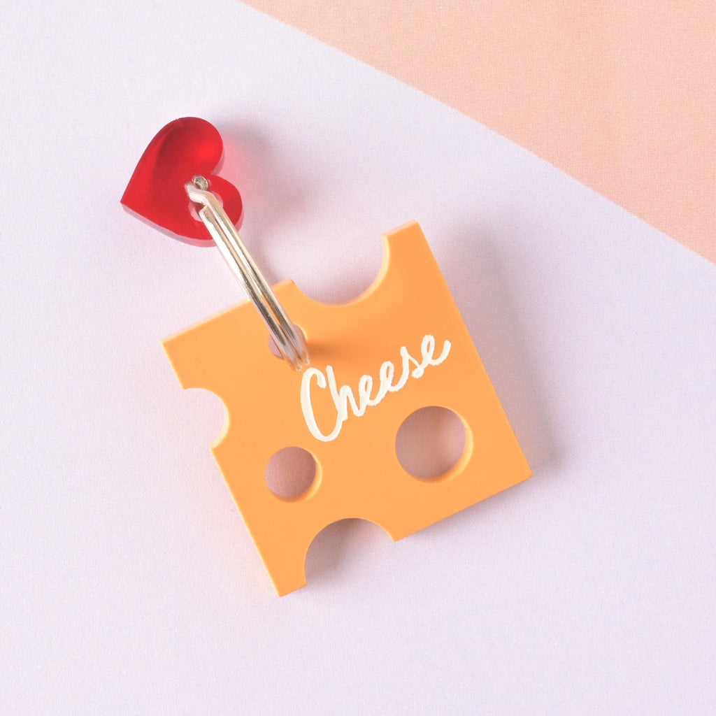 A custom pet ID tag shaped like a slice of Swiss cheese (orange acrylic) with "holes" cut out. The tag is engraved with the word "Cheese" and accented with a small translucent red heart charm, attached to a split ring. Personalized Cheese Pet Tag - Custom Tag for Cheese Lovers, dogs and cats - Engraved ID Name and Contact Info Included