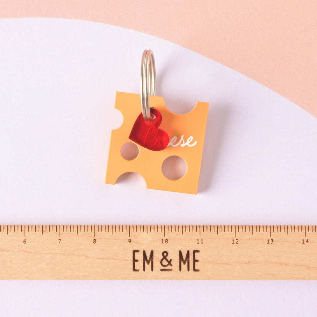 A custom pet ID tag shaped like a slice of Swiss cheese (orange acrylic) with "holes" cut out. The tag is engraved with the text "Cheese" and accented with a small red heart charm, shown above a wooden ruler for size reference. Personalized Cheese Pet Tag - Custom Tag for Cheese Lovers, dogs and cats - Engraved ID Name and Contact Info Included
