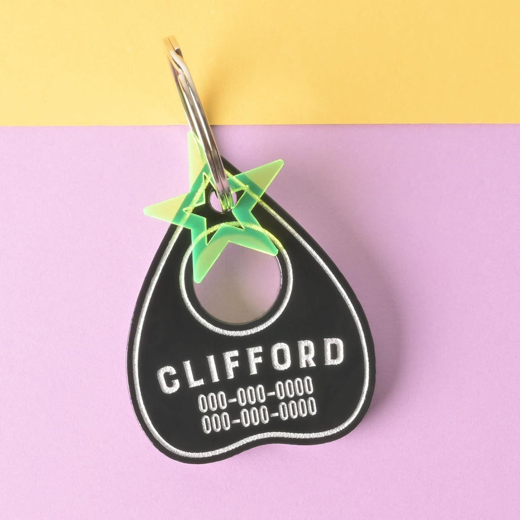 A personalized black Ouija planchette-shaped pet tag with the name "CLIFFORD" and a placeholder phone number on it. A small, translucent green star charm is attached. The tag is connected to a silver ring and is displayed on a background that is half solid yellow and half solid purple.