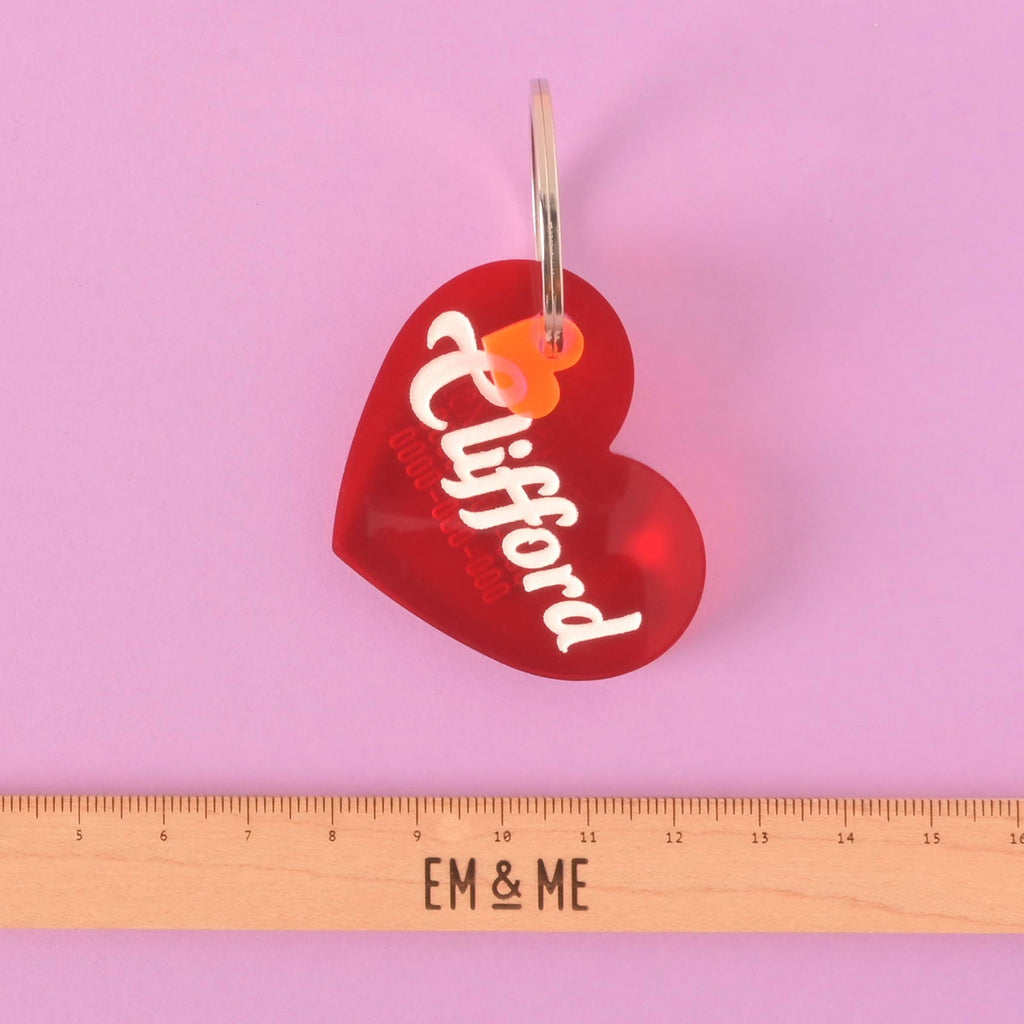 A personalized red heart-shaped pet tag with the name "Clifford" and a placeholder phone number on it. A small orange heart charm is attached. A wooden ruler with the "EM & ME" logo is placed below the tag to show its size. The items are shown on a solid purple background.