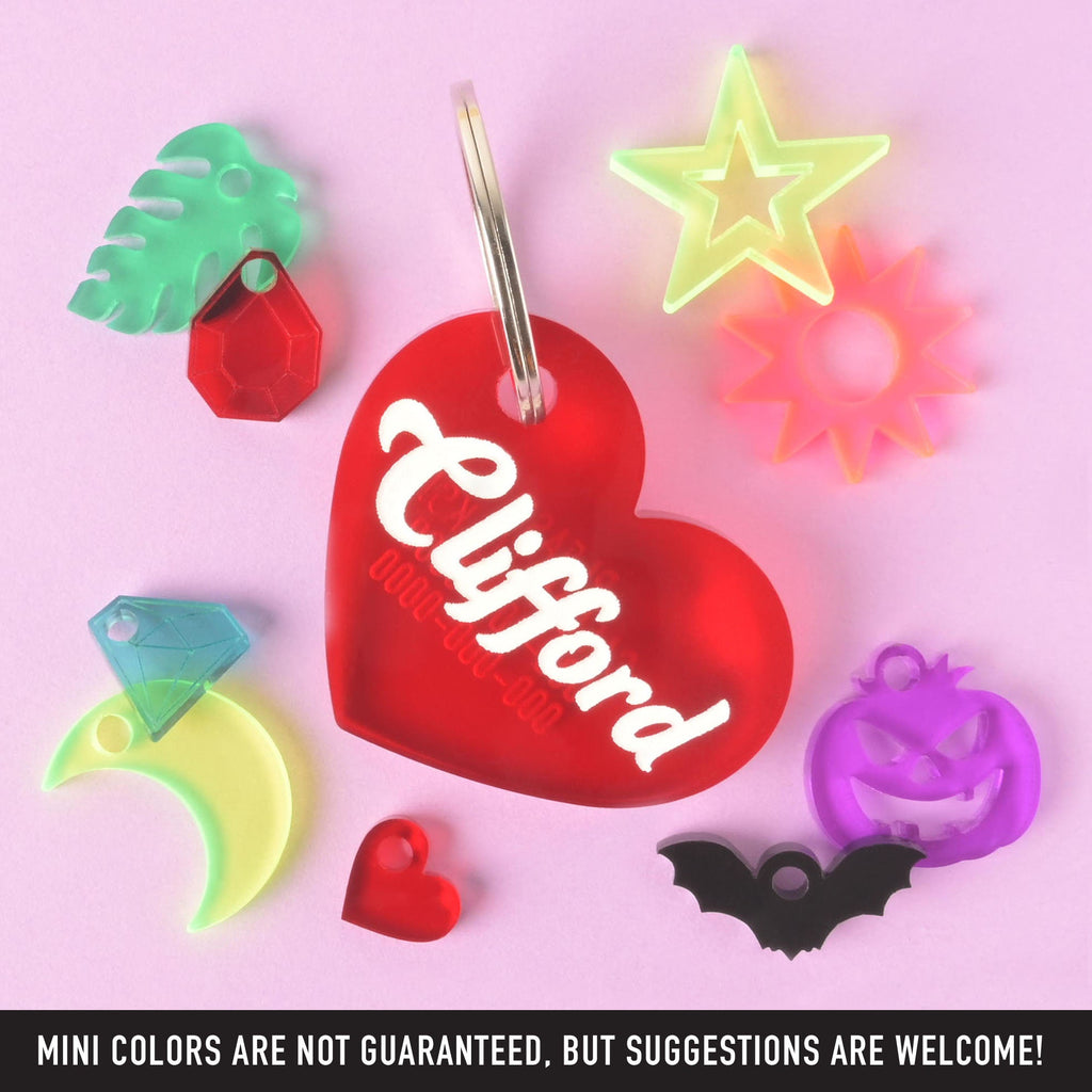 A collection of pet tags and charms scattered on a solid pink background. The central piece is a personalized red heart-shaped pet tag with the name "Clifford" and a placeholder phone number on it. Surrounding it are various charms, including a green monstera leaf, a red gemstone, a yellow crescent moon with a blue diamond, a purple carved pumpkin, a black bat, a small red heart, a neon yellow star, and a pink and orange sunburst. The image also contains the text "MINI COLORS ARE NOT GUARANTEED, BUT SUGGEST