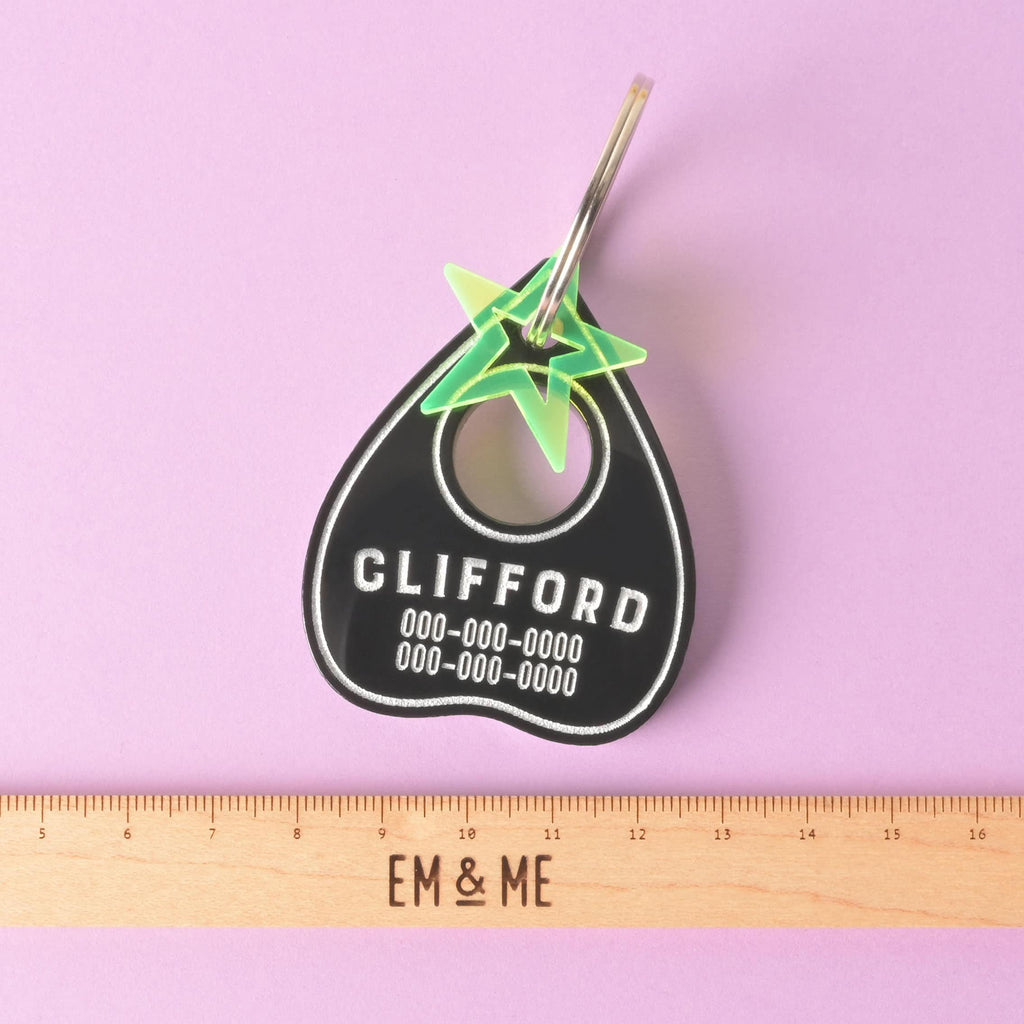 A black Ouija planchette-shaped pet tag with the name "CLIFFORD" and a placeholder phone number on it. A small, translucent green star charm is attached. A wooden ruler with the "EM & ME" logo is placed below the tag to show its size. The items are shown on a solid purple background.