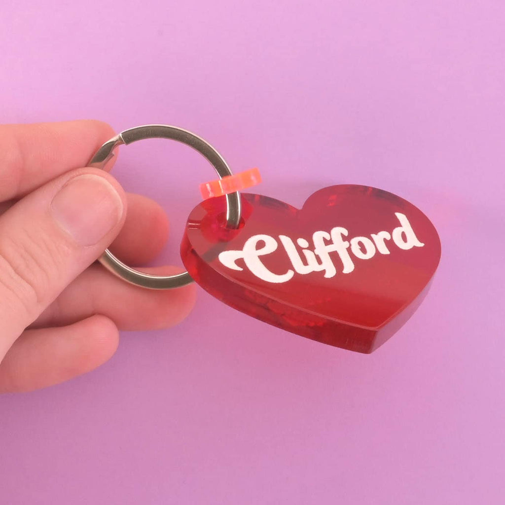 A person's hand holds a personalized red heart-shaped pet tag. The tag has the name "Clifford" and a small orange heart charm attached. It's connected to a silver ring, perfect for a collar, and is shown against a solid purple background.