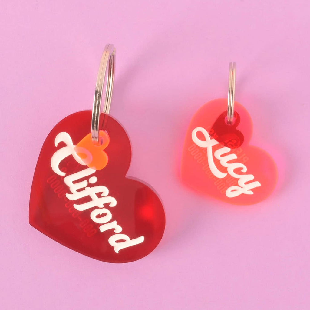 A pair of personalized heart-shaped pet tags for "Clifford" and "Lucy." The larger, red tag has the name "Clifford" and a small orange heart charm. The smaller, pink tag has the name "Lucy" and a small red heart charm. Both tags have a placeholder phone number and are attached to a silver ring for a collar. The items are displayed on a solid pink and solid purple background.