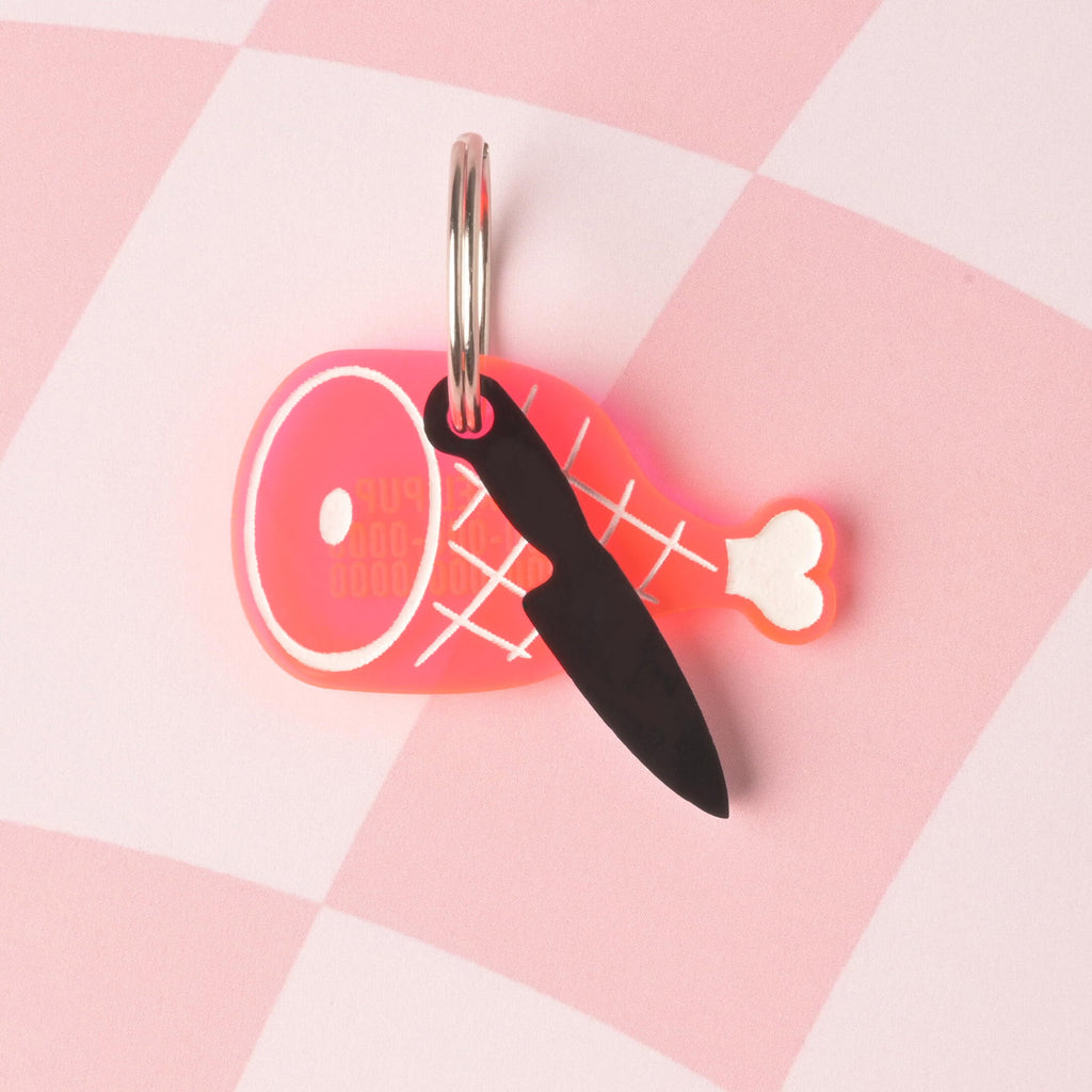 fake knife, Seasonal Mini,  Tiny Accessory for Pet ID Tag, Cats and Dogs Add-on Charm A personalized pet ID tag shaped like a pink ham/roast with white engraving. A small, black opaque acrylic knife charm hangs from the same ring. The tags are displayed on a pink checkered background.