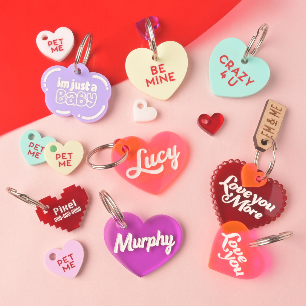 A large assortment of heart-shaped acrylic pet ID tags in red, pink, purple, mint, and cream. Tags feature slogans like 'Be Mine,' 'Crazy 4 U,' 'I'm Just a Baby,' 'Love you More,' and personalized names like 'Lucy' and 'Murphy.' The collection also includes a pixelated red heart tag and several small 'Pet Me' charms. Small Pet Me Heart, Candy Inspired, Valentine's Day, Seasonal Mini, Tiny Accessory for Pet ID Tag, Cats and Dogs Add-on Charm