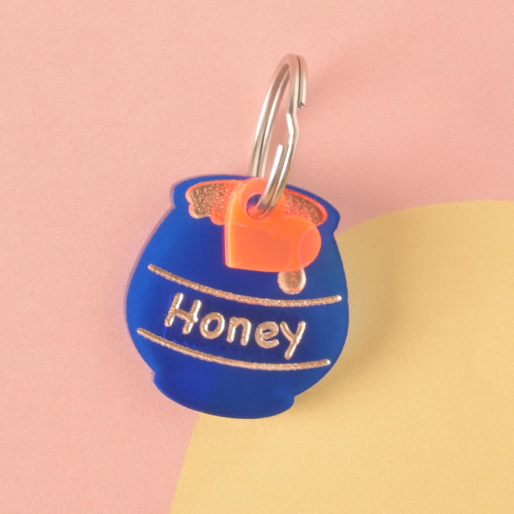 A personalized, matte blue acrylic pet ID tag shaped like a honey pot, engraved with the name 'Honey' and a gold drip graphic. The tag is accented with a small orange heart charm.