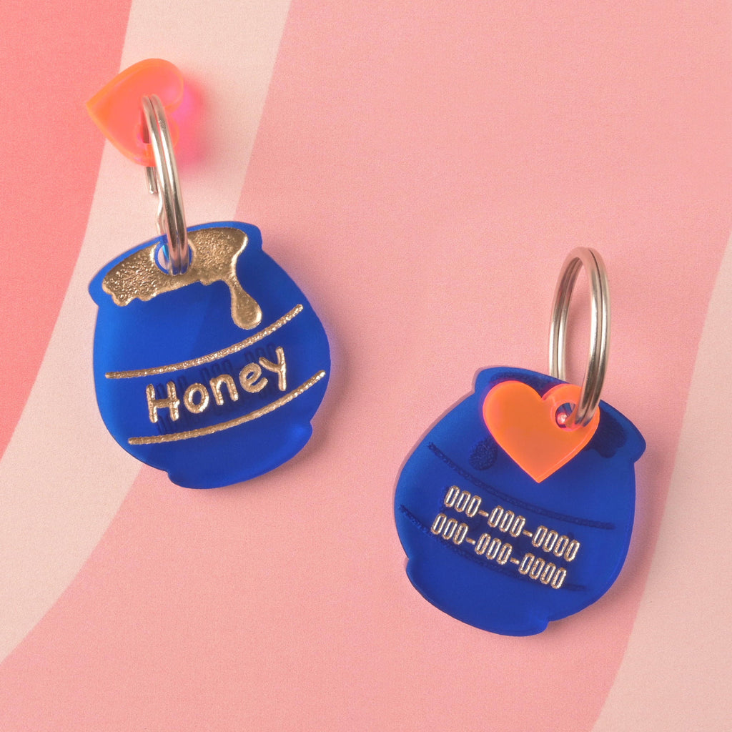 Two sides of a custom, matte blue acrylic pet ID tag shaped like a honey pot. The front is engraved with 'Honey' and a gold drip graphic, and the back is engraved with phone numbers. The tag is accented with a small orange heart charm.