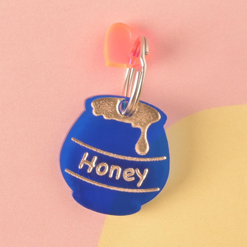 A personalized, matte blue acrylic pet ID tag shaped like a honey pot, engraved with the name 'Honey' and a gold drip graphic. The tag is accented with a small pink/orange heart charm.
