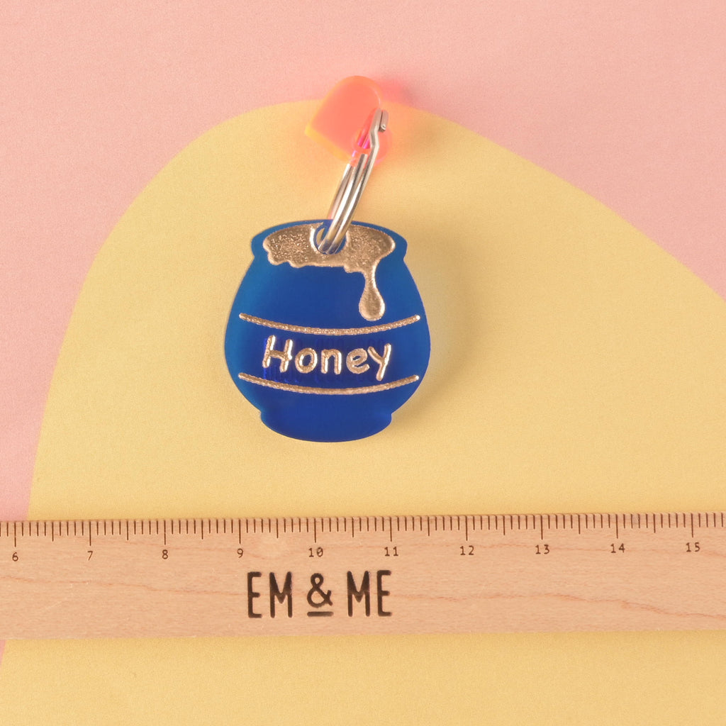 A personalized, matte blue acrylic pet ID tag shaped like a honey pot, engraved with 'Honey' and a gold drip graphic. The tag is accented with a small pink/orange heart charm and shown above a wooden ruler for size reference.