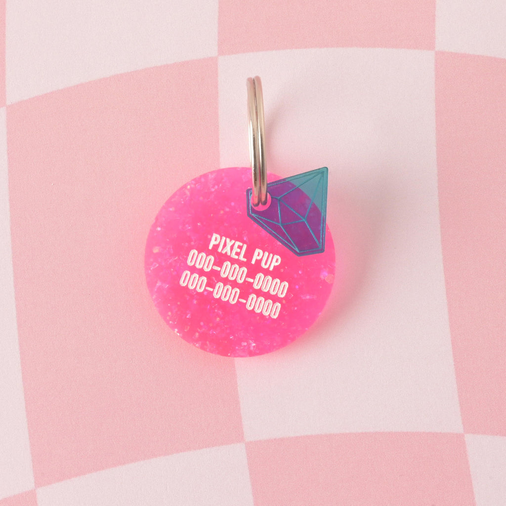 A custom, circular pet ID tag made from vibrant hot pink glitter acrylic. The tag is engraved with the name 'PIXEL PUP' and three lines of phone numbers, and accented with a small blue diamond-shaped gemstone charm with iridescent details. Material Babe, Barbie Inspired Personalized Pet Tag, ID Tag for Cats and Dogs, Limited Edition, Custom Name and Contact Info Engraved