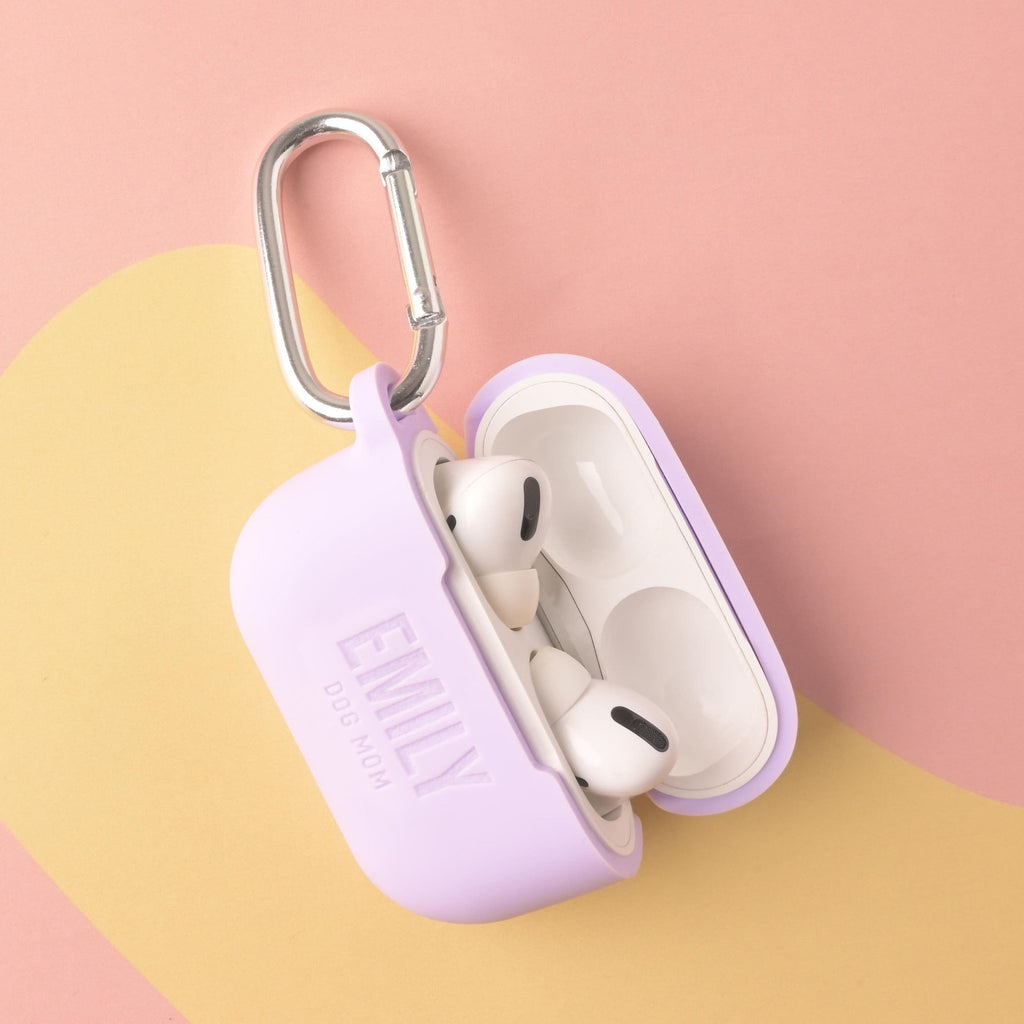 A personalized silicone AirPods Pro case in a lavender or light purple color, open to show the earbuds inside. The case is custom engraved with the text 'EMILY' and 'DOG MOM' and features a silver metal carabiner clip.