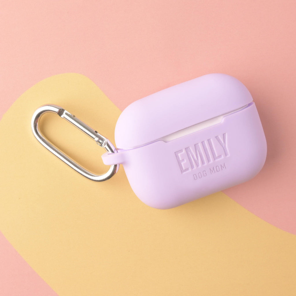 A personalized silicone AirPods Pro case in a solid lavender color, custom engraved with the text 'EMILY' and 'DOG MOM,' and featuring a large silver metal carabiner clip.
