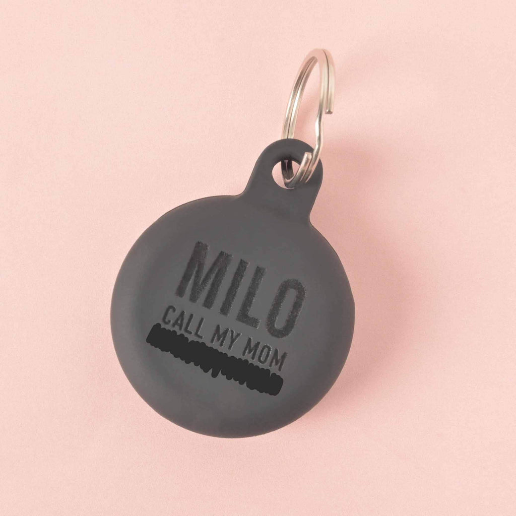 A personalized black Silicone round pet tag that says "MILO" and "CALL MY MOM" with a placeholder phone number on it. The tag is attached to a silver ring, perfect for a collar, and is displayed on a solid pink background.