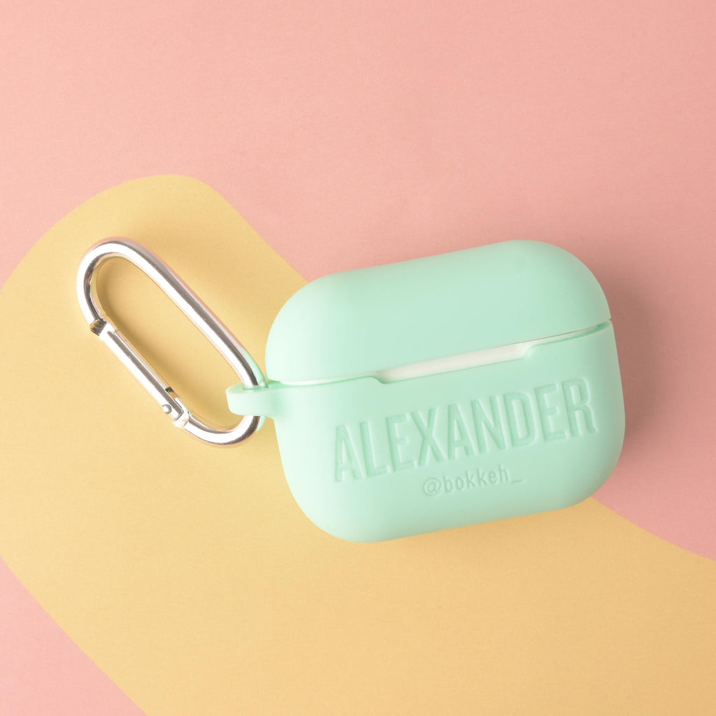 A personalized silicone AirPods Pro case in a mint green color, custom engraved with the name 'ALEXANDER' and the social media handle '@bokkeh_'. The case is attached to a large silver metal carabiner clip.