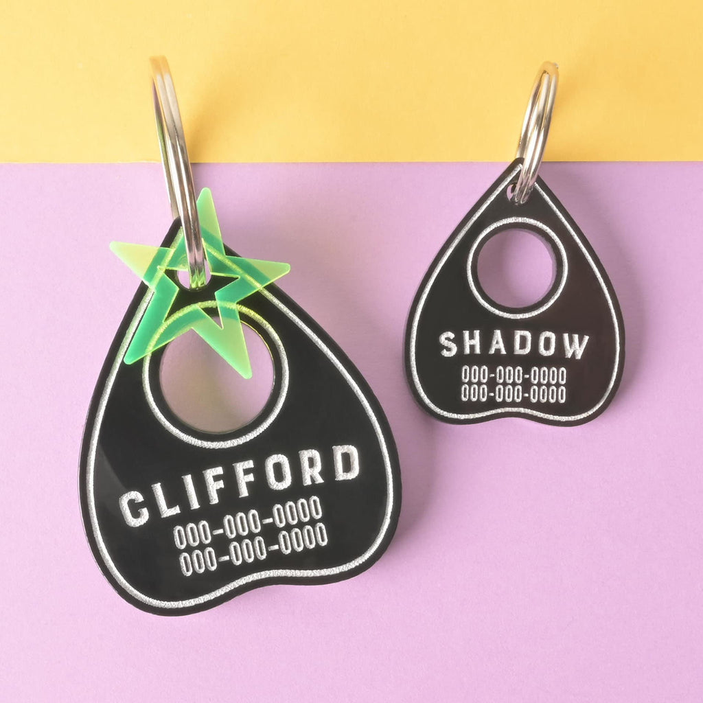 A pair of personalized black Ouija planchette-shaped pet tags in two sizes. The larger tag has the name "CLIFFORD" and a translucent green star charm. The smaller tag has the name "SHADOW." Both tags have a placeholder phone number and are attached to a silver ring for a collar. The items are displayed on a background that is half solid yellow and half solid purple.
