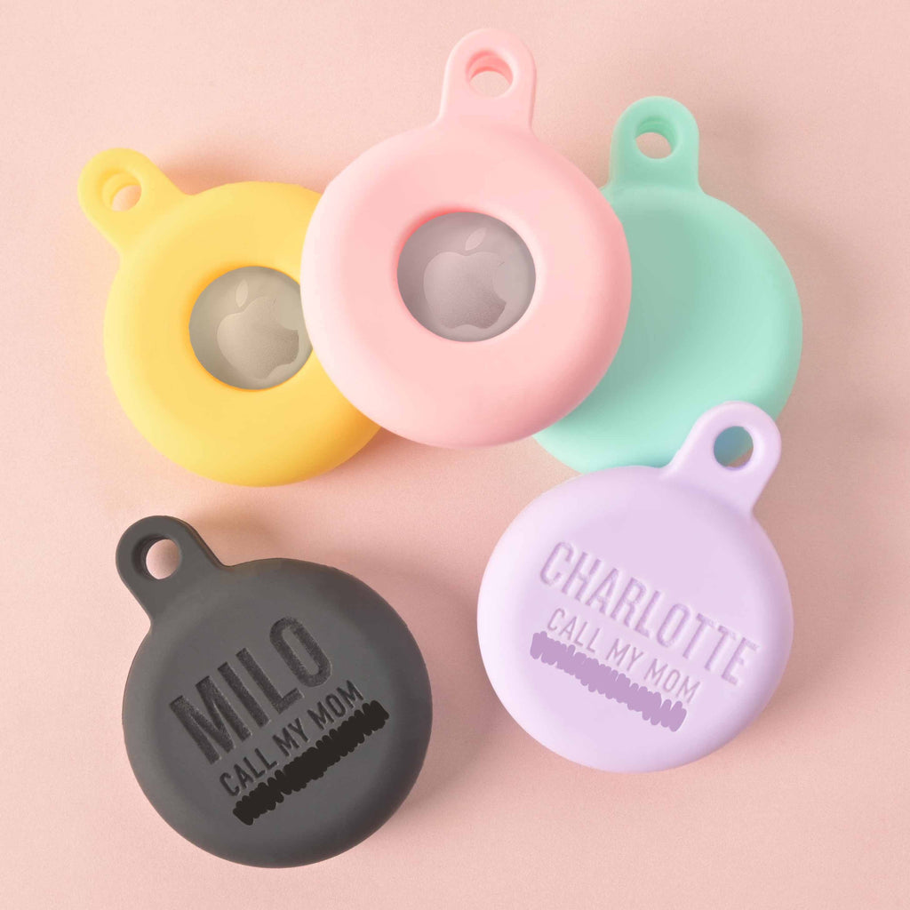 A set of five personalized AirTag holders for pets, each in a different color. The holders include a yellow one, a pink one, a mint green one, a gray one that says "MILO," and a light purple one that says "CHARLOTTE." The holders are displayed on a solid pink background.