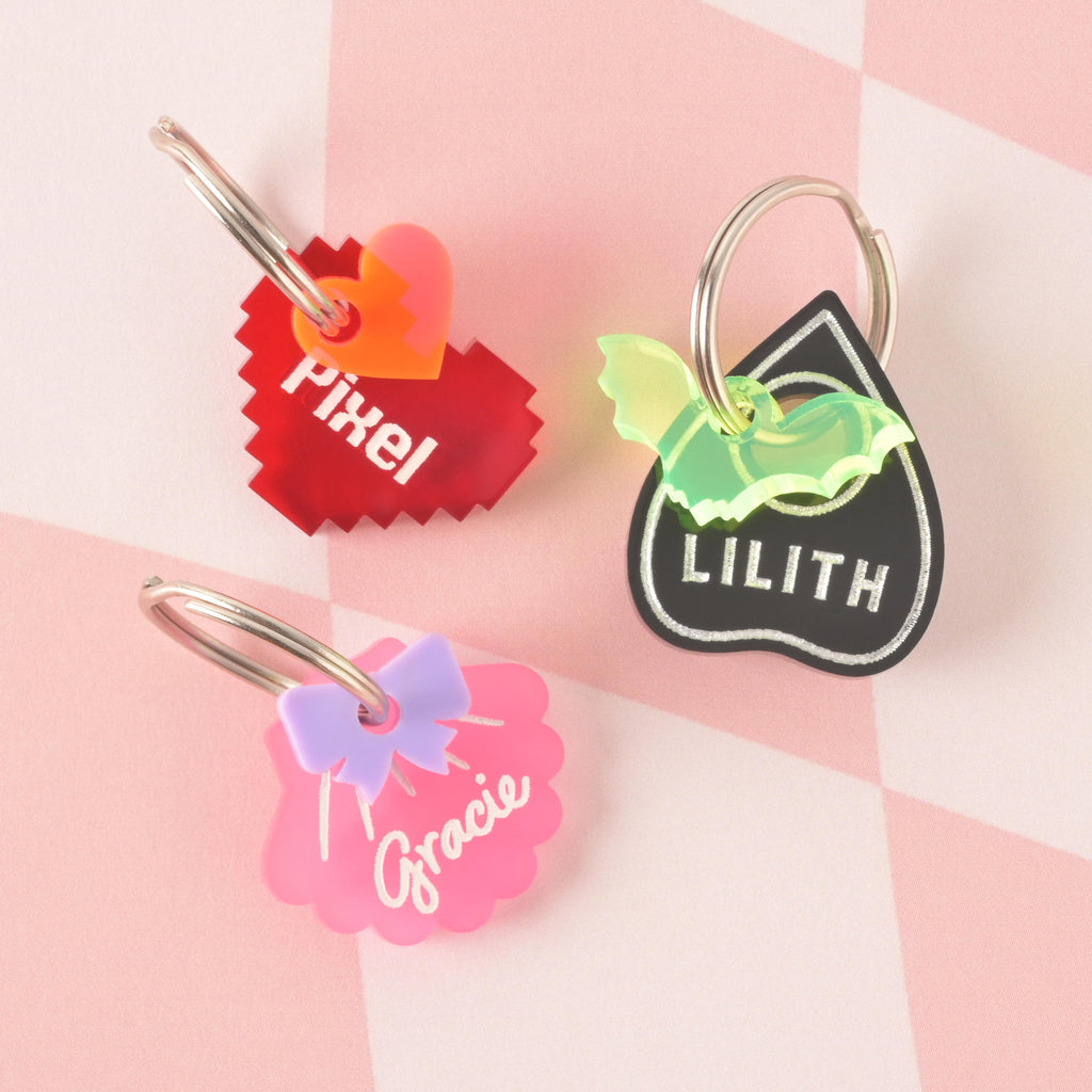 three personalized pet tags. One is a red pixelated heart with the name "Pixel" and a small orange heart, one is a black Ouija board planchette with the name "LILITH" and a translucent winged symbol, and one is a pink seashell with the name "Gracie" and a purple bow. All are secured with a silver ring for easy attachment to a collar and are displayed on a pink and white checkered background.