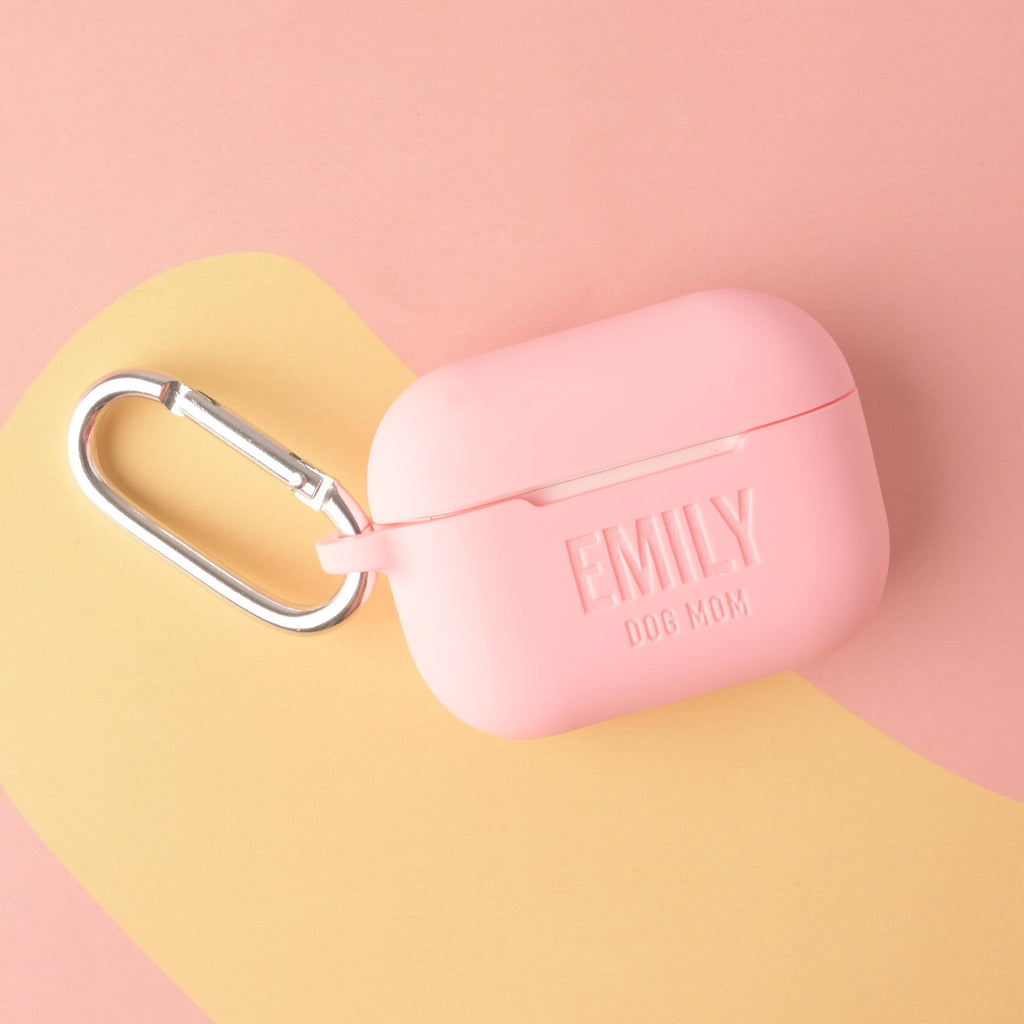 A personalized silicone AirPods Pro case in baby pink, custom engraved with the text 'EMILY' and 'DOG MOM,' and featuring a large silver metal carabiner clip.