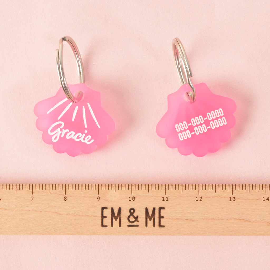 pair of pink seashell-shaped pet tags shown with a ruler for scale. The front of one tag says "Gracie," while the back of the other has a placeholder phone number. The tags are resting on a solid pink background with a ruler below them that reads "EM & ME."