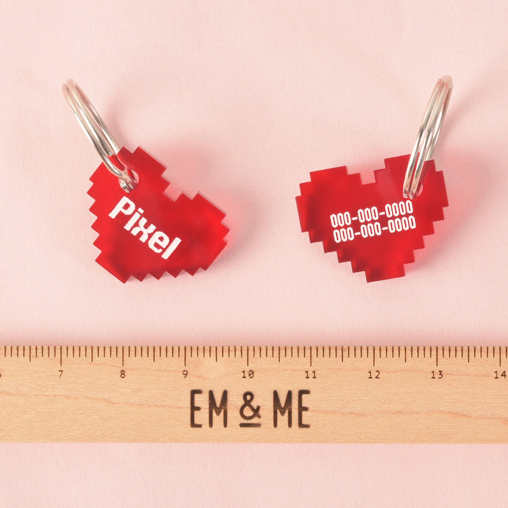 A red pixelated heart pet ID tag with a silver ring. The front of the tag says "Pixel," and the back shows a placeholder phone number. The tag is placed next to a ruler with the "EM & ME" logo to show its size. The background is a solid pink color.
