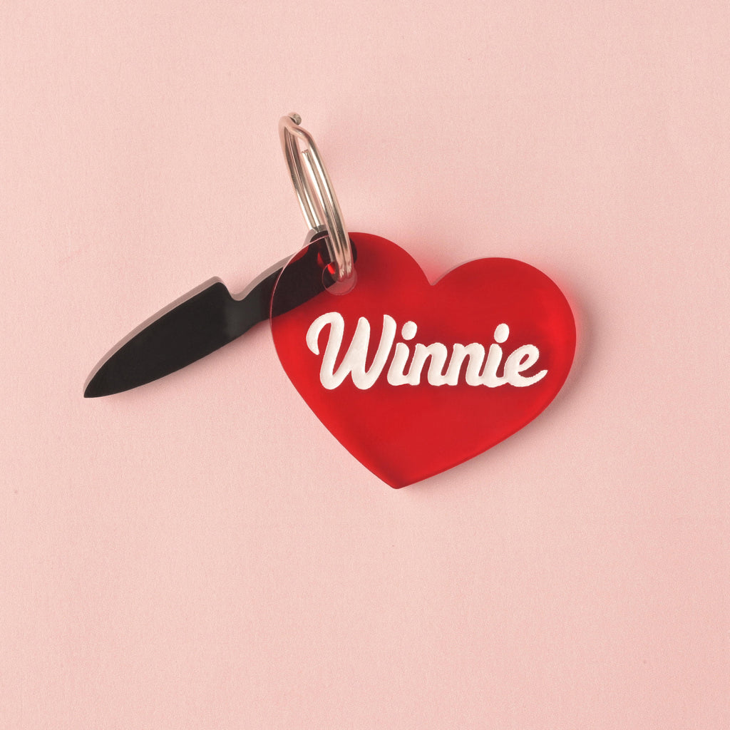 A personalized, translucent red acrylic heart-shaped pet ID tag, engraved with the name 'Winnie' in white script. A small, black opaque acrylic knife charm hangs from the same ring. fake knife, Seasonal Mini,  Tiny Accessory for Pet ID Tag, Cats and Dogs Add-on Charm
