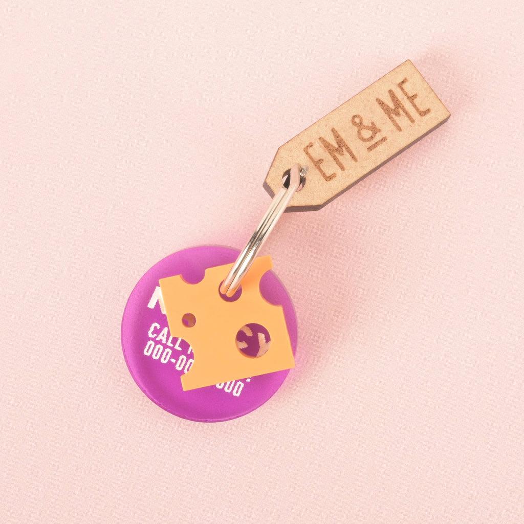 A purple round pet tag with a placeholder phone number and a small, orange cheese charm attached. The tags are connected to a silver ring, with a small rectangular wooden charm that reads "EM & ME." The items are shown on a light pink background.