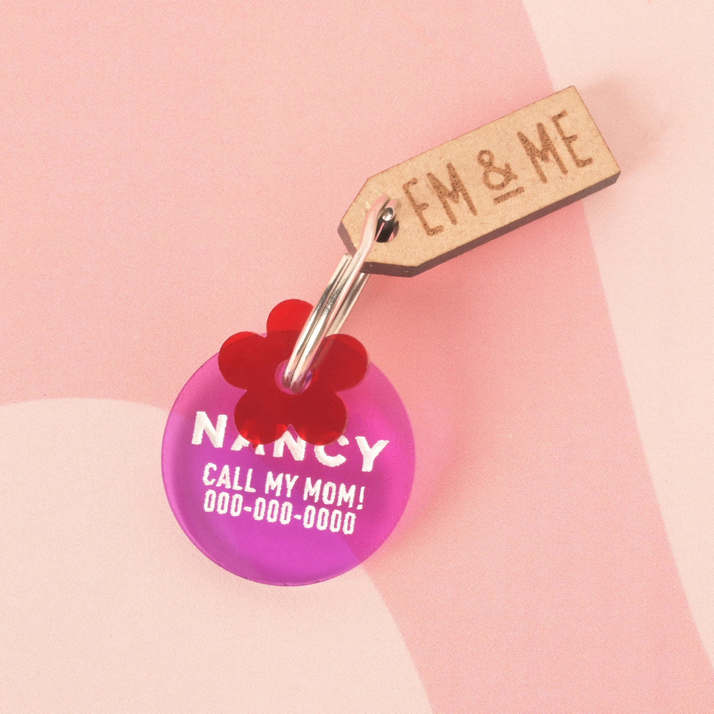 A round, purple transparent pet tag with the name "NANCY" and a placeholder phone number. A small red flower charm is attached to the tag, along with a rectangular wooden charm that reads "EM & ME." The items are connected to a silver ring and are shown on a light pink and white checkered background.
