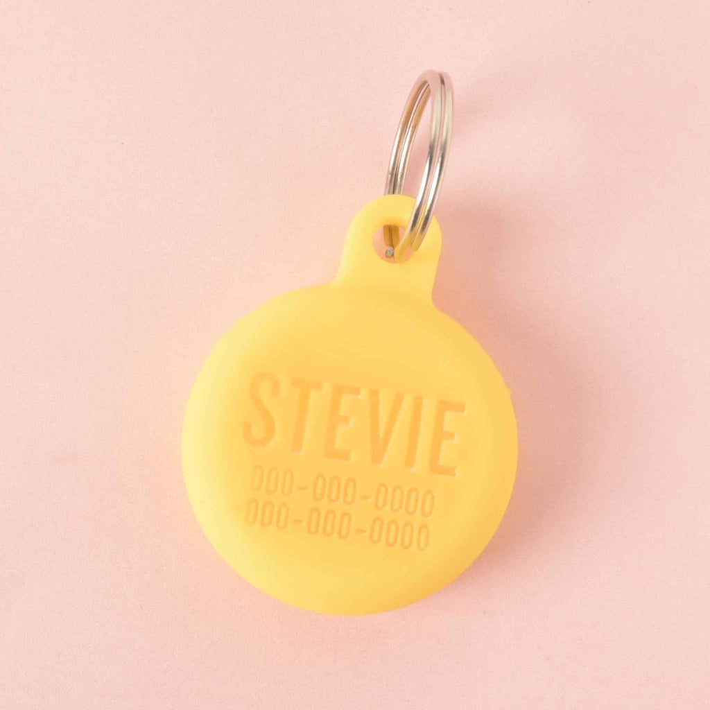 A personalized yellow round pet tag that says "STEVIE" and has a placeholder phone number on it. The tag is attached to a silver ring, perfect for a collar, and is displayed on a light pink background.