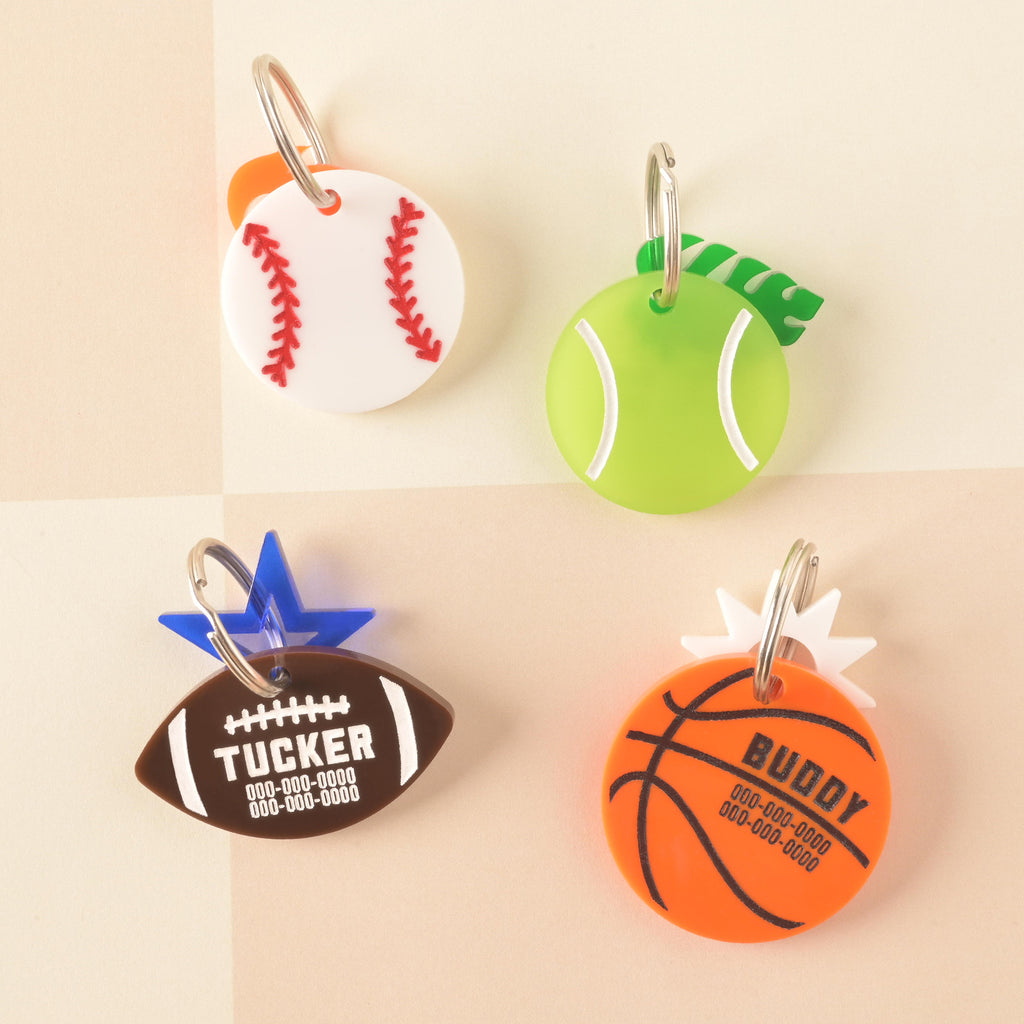 A set of four custom, acrylic pet ID tags shaped like sports balls: a white baseball with red stitching, a neon green tennis ball with a green leaf accent, a brown football for 'TUCKER' with a blue star, and an orange basketball for 'BUDDY' with a white sunburst charm. Tennis Ball Pet Tag, Personalized Pet tag, Cat or Dog ID Tag, summer ball tag