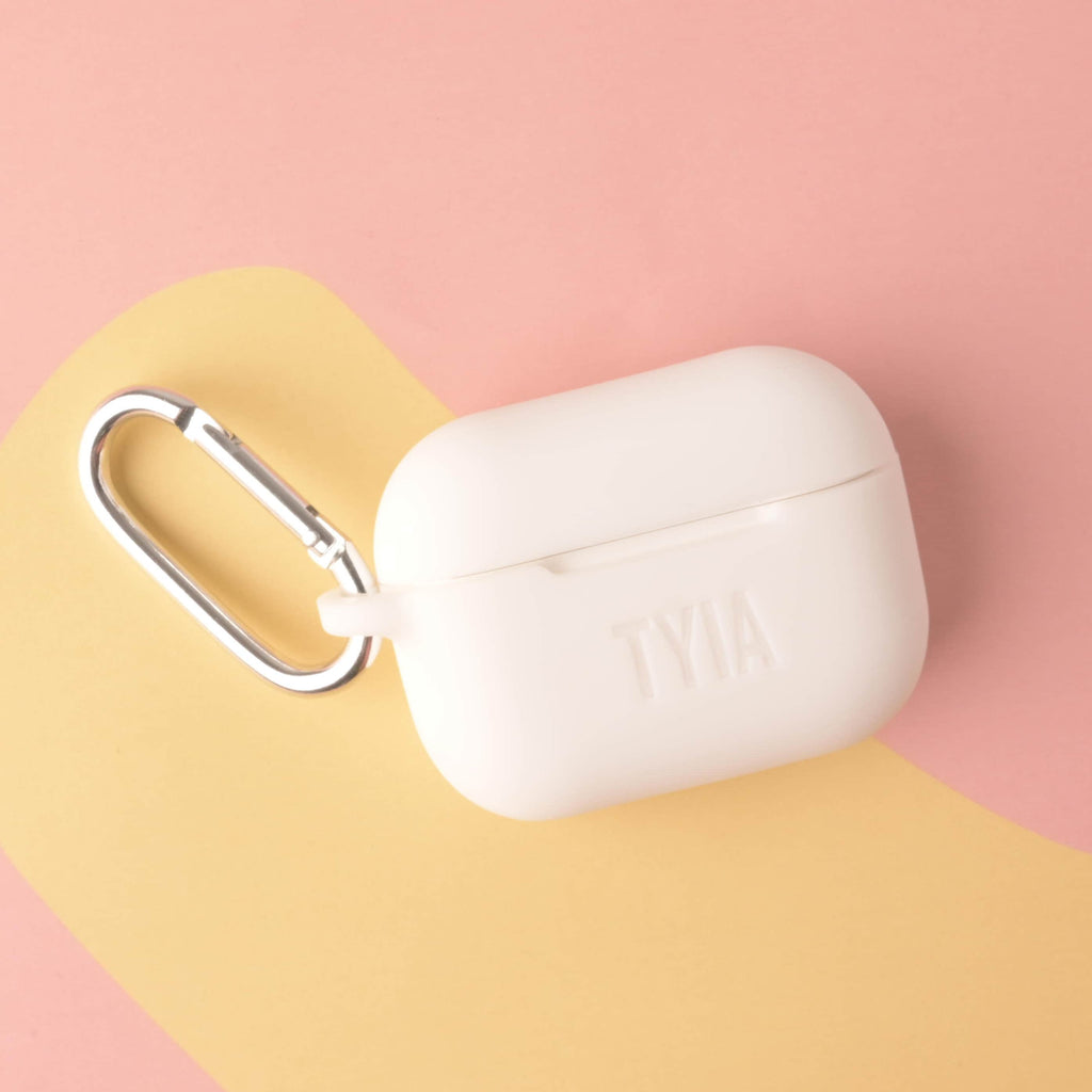 personalized silicone AirPods Pro case in a soft white or cream color, custom engraved with the name 'TYIA,' and featuring a large silver metal carabiner clip.