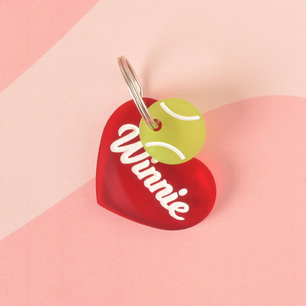 A personalized red heart-shaped pet tag with the name "Winnie" written in white. A yellow-green tennis ball charm is attached on top of the tag. Both are connected to a silver ring, perfect for a collar, and are displayed on a light pink and solid pink background with a wavy design.