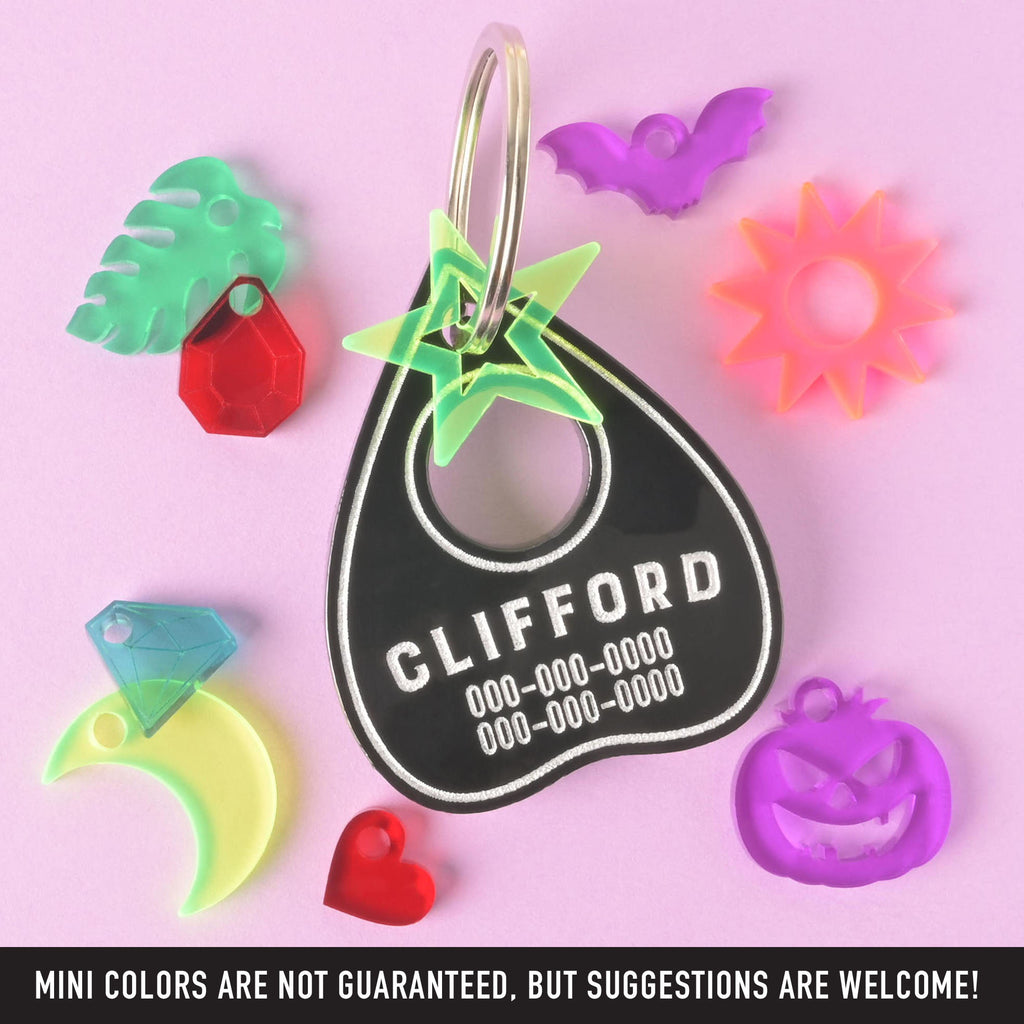 A collection of pet tags and charms scattered on a solid purple background. The central piece is a personalized black Ouija planchette-shaped pet tag with the name "CLIFFORD" and a placeholder phone number on it. Surrounding it are various charms, including a green monstera leaf, a red gemstone, a yellow moon, a blue diamond, a red heart, a purple bat, an orange sunburst, a purple carved pumpkin, and a green star.