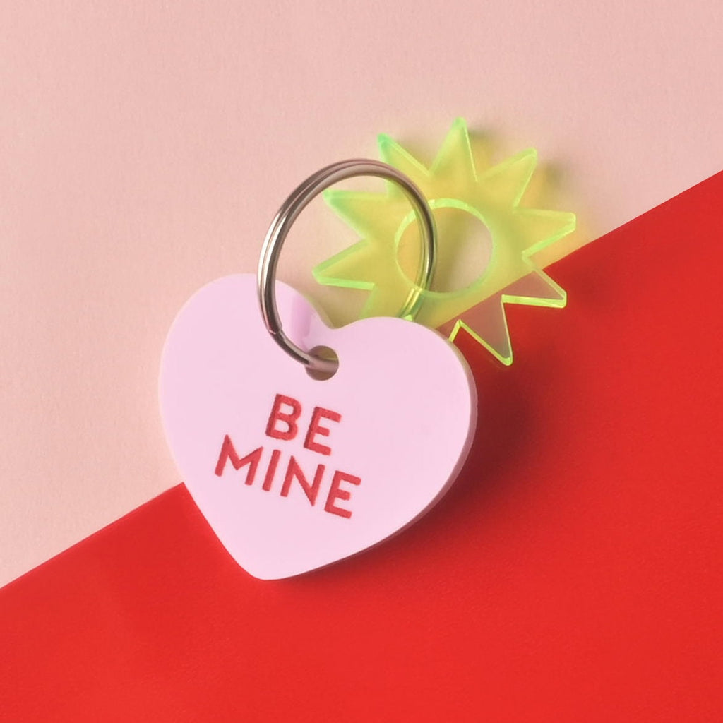 Close-up of a single light pink heart-shaped acrylic pet ID tag, engraved with the red slogan 'BE MINE.' A neon yellow-green sun/starburst charm is attached to the silver split ring. The tag is displayed on a color-blocked background of light pink and bright red. Conversation Hearts, Valentines Heart Dog tag, Customize front and back, ID tag for cats and dogs, Personalized Pet Tag
